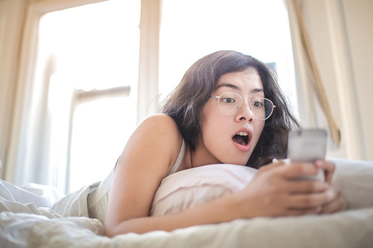 girl with black hair and glasses looking at phone in bed with a shocked look