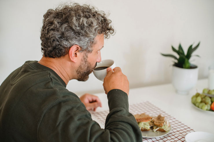 man with grey hair sat at table drinking coffee and eating a sandwich