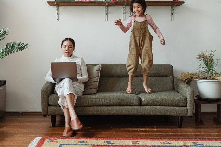 mother trying to work on laptop sat on couch with daughter jumping on it next to her