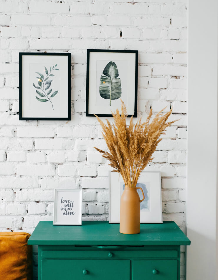framed leaf prints inblack frames on white brick wall with orange vase and green drawers