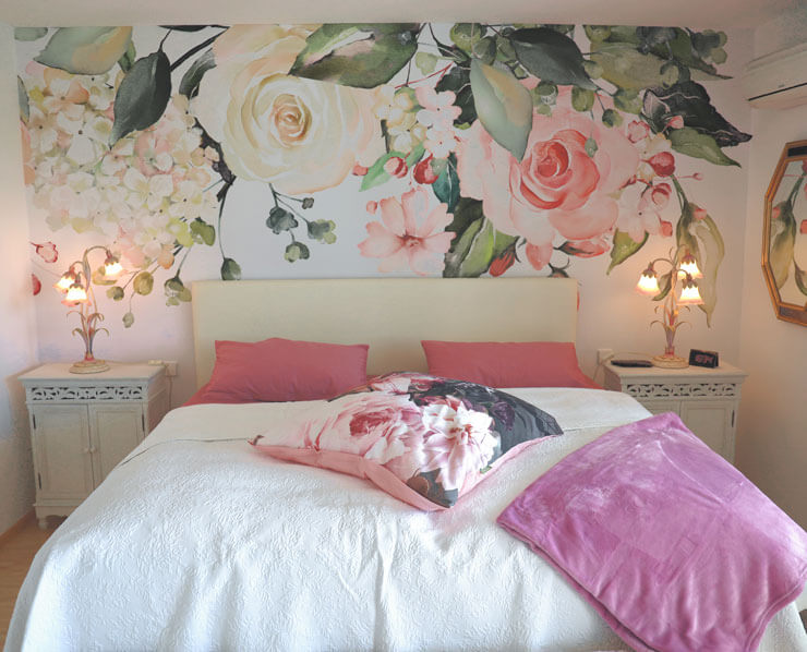 White and pink bedroom with white bedsheets, pink cushions and a long pink floral wallpaper over the bed
