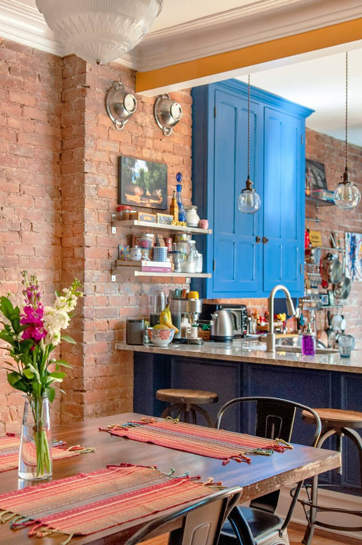 Bare red brick walls in a kitchen with light blue cupboards and a pine dining table