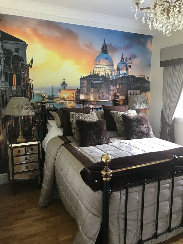 venice sunset wall mural behind bed in bedroom with faux fur cushions and glass chandelier